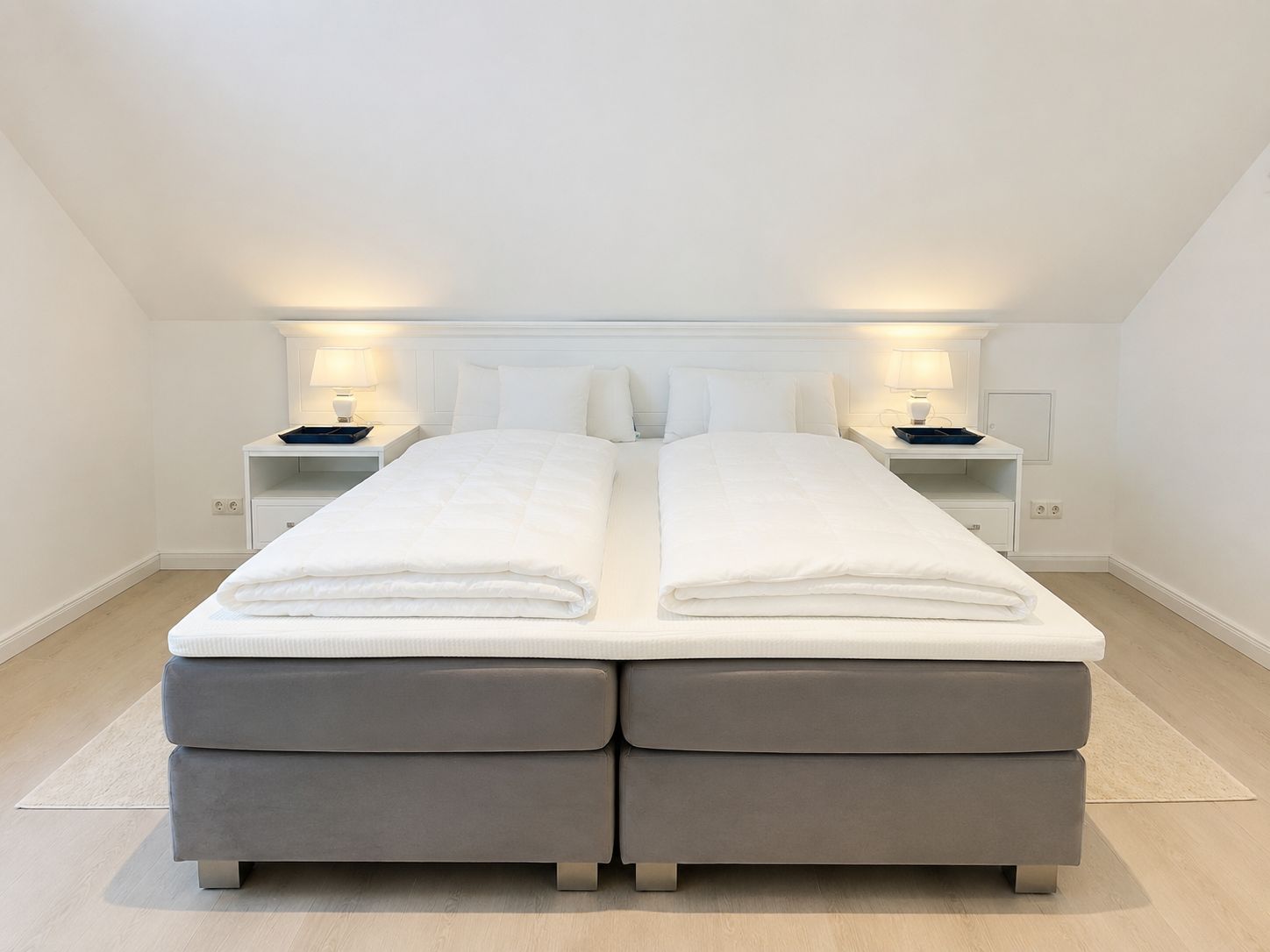 Ein Doppelbett mit grauem Polsterfuß, weißer Bettwäsche und zwei Nachttischen mit Lampen.