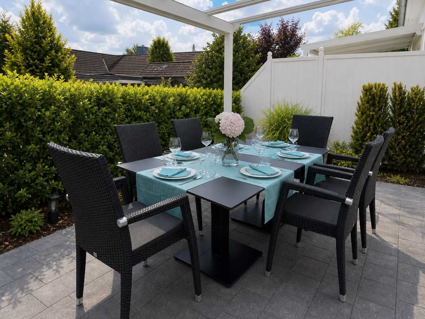 Eine Terrasse im Freien mit einem für sechs Personen gedeckten Tisch, umgeben von Korbstühlen, unter einer weißen Pergola.