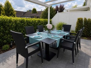 Een terras buiten met een tafel gedekt voor zes personen, omringd door rieten stoelen, onder een witte pergola.
