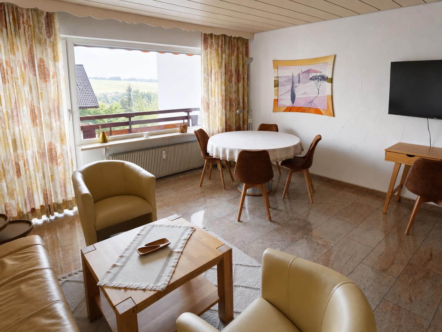 Ein Wohnzimmer mit rundem Tisch, Stühlen, einer Couch, einem Fernseher und einem Fenster mit Vorhängen.