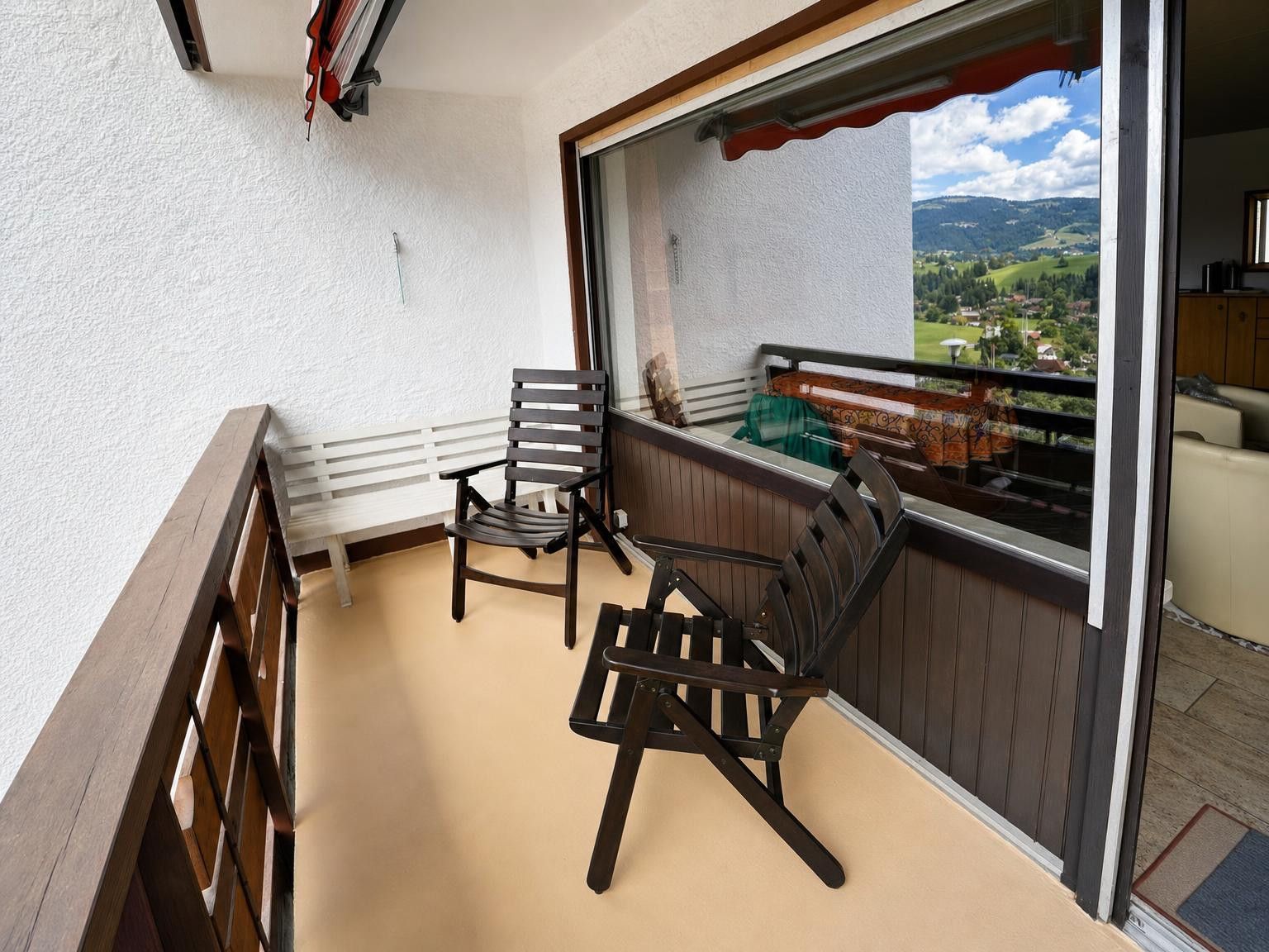 Ein Balkon mit zwei Holzstühlen und einer weißen Sitzbank, mit Blick auf eine malerische Landschaft.