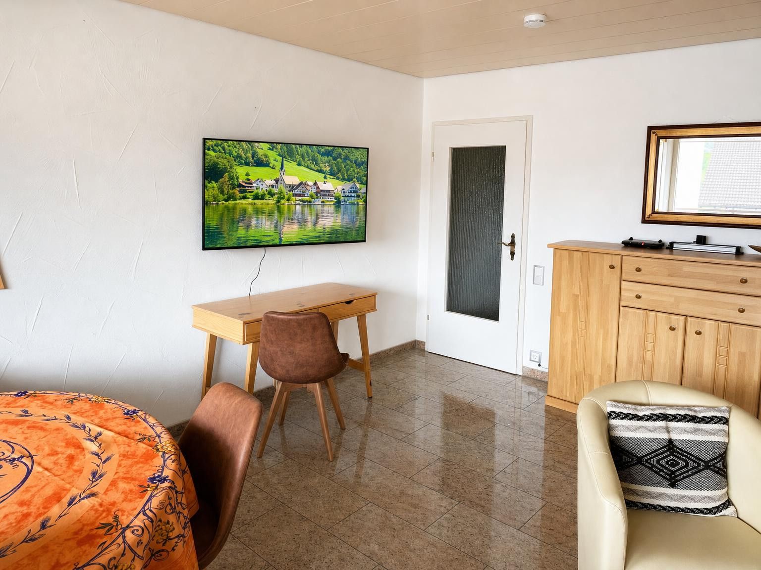 Ein Zimmer mit einem Holztisch, einem Stuhl, einem Bett mit gemustertem Überwurf, einem Fernseher an der Wand und einem Schrank.