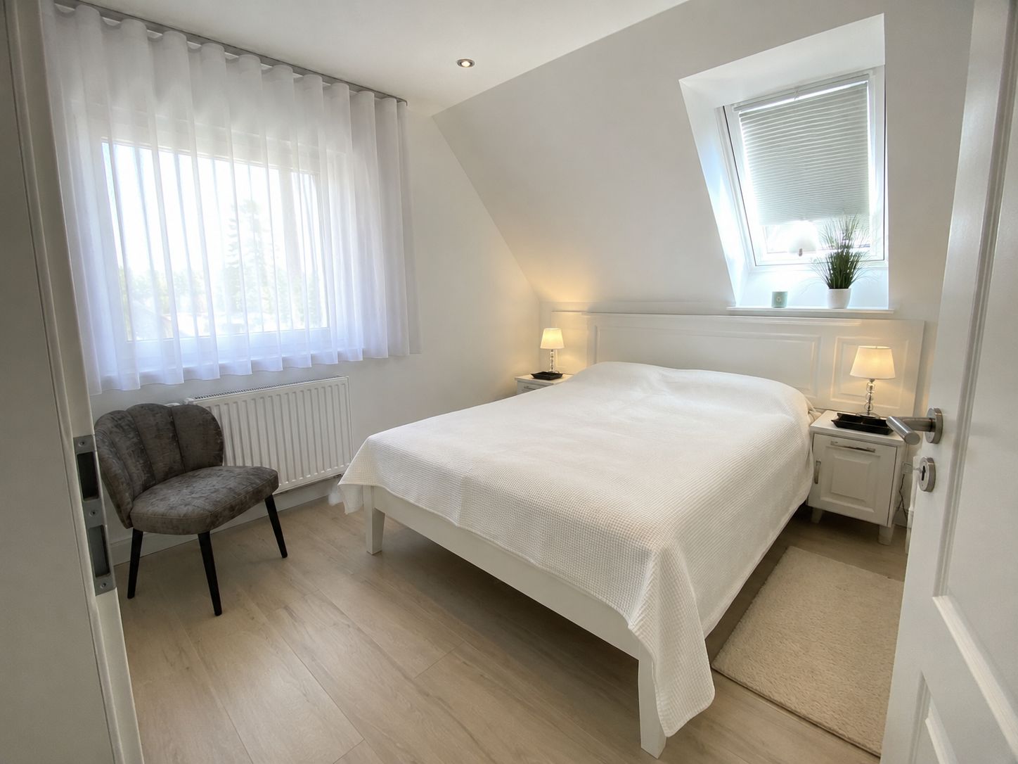 Ein Schlafzimmer mit einem Bett, einem Sessel, einem Fenster mit Jalousien und einem Nachttisch mit Lampe.