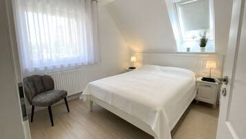 Ein Schlafzimmer mit einem Bett, einem Sessel, einem Fenster mit Jalousien und einem Nachttisch mit Lampe.
