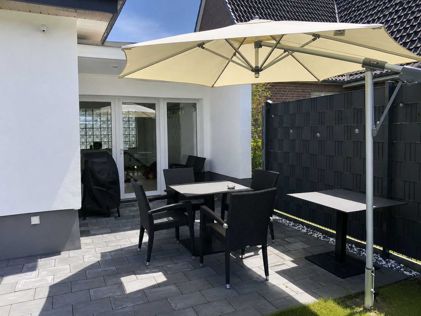 An outdoor patio with a table and chairs, a large umbrella, and a view of a house through glass doors.