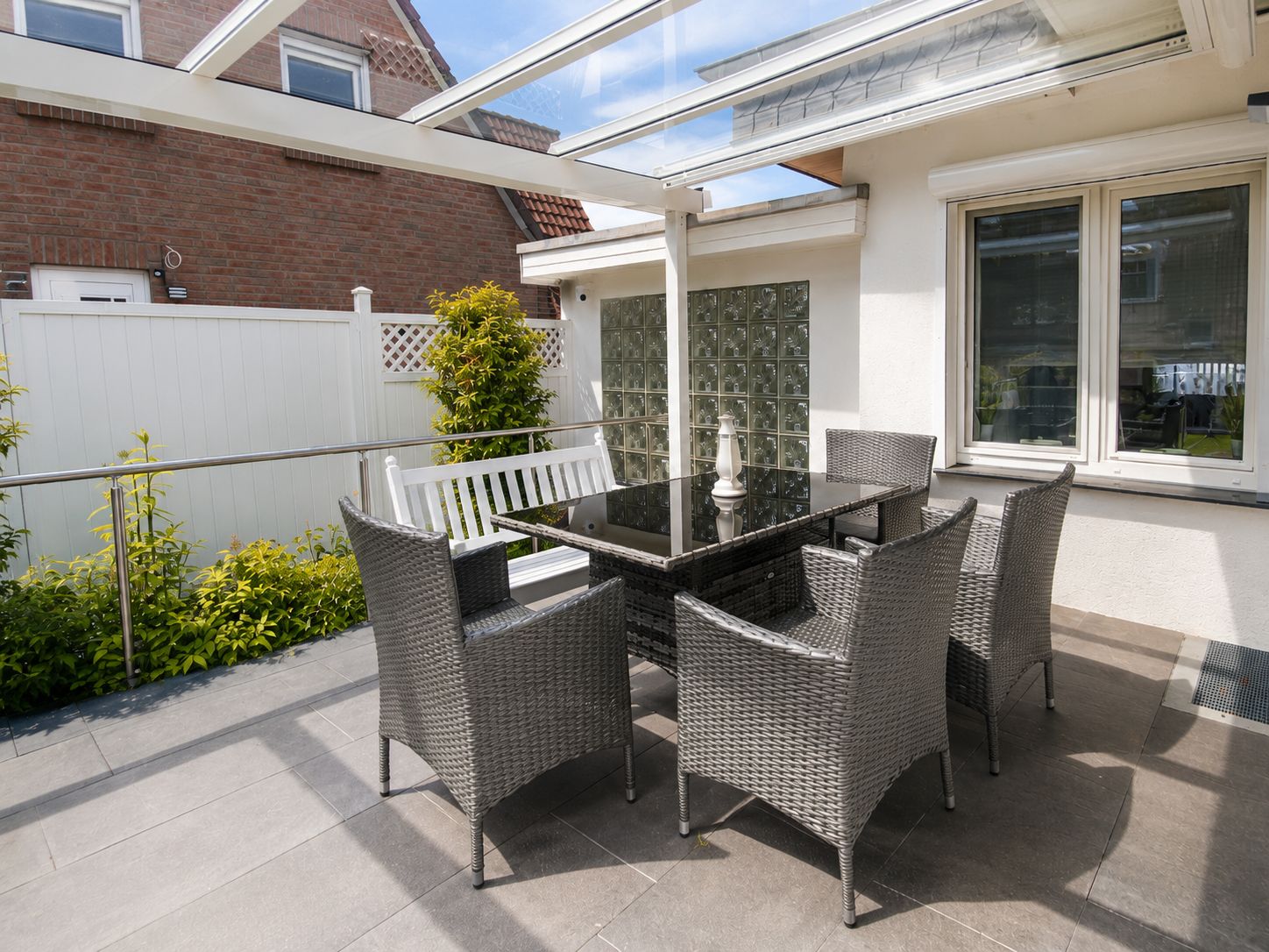 Eine Terrasse im Freien mit einem Glastisch und Korbstühlen, umgeben von einem weißen Zaun und Pflanzen.