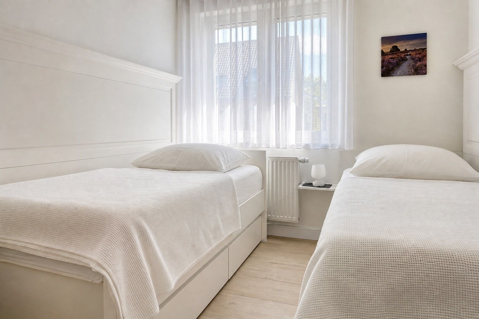 Ein Hotelzimmer mit zwei Betten, einem Fenster mit durchsichtigen Vorhängen, einem Heizkörper und einem kleinen Tisch mit einer Lampe.