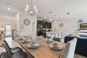 A modern dining area with a long table set for six, a kitchen in the background, and a living area with a sofa and TV. - ChampionsGate Townhome w/ Private Pool (Kissimmee)