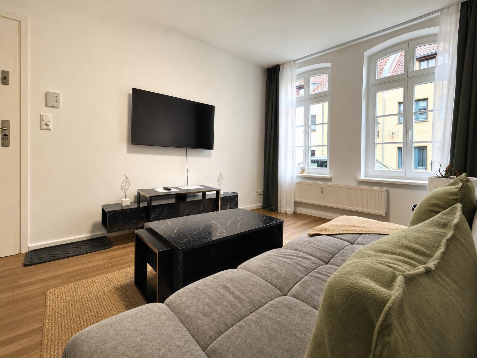 Ein modernes Wohnzimmer mit einem an der Wand befestigten Flachbildfernseher, einem schwarzen Couchtisch, einem grauen Sofa und einem Fenster mit Vorhängen.