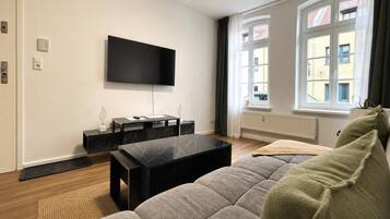 Een moderne woonkamer met een flatscreen televisie aan de muur, een zwarte salontafel, een grijze bank en een raam met gordijnen.