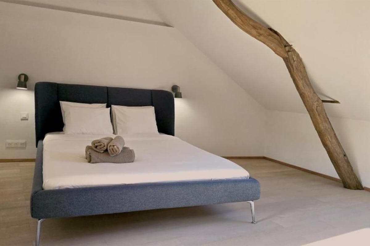 A modern bedroom with a sloped ceiling, a bed with a dark headboard, and a wooden beam structure.