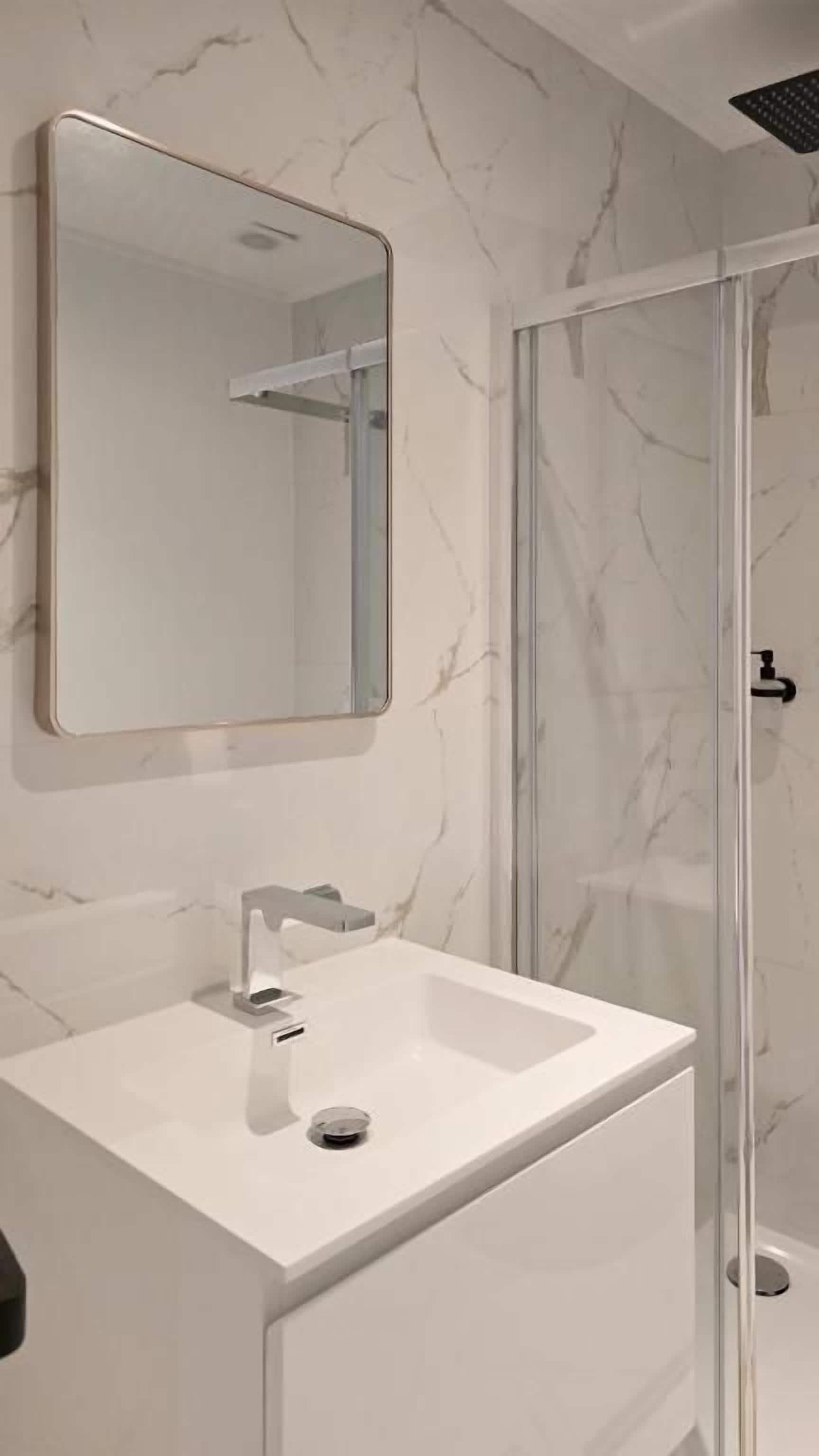 Un baño con pared de mármol blanco, un espejo rectangular, un lavamanos blanco y un box de ducha de vidrio.