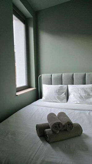 A neatly made bed with white linens and a grey headboard, a window on the left side, and a pair of rolled towels on the bed. - botanique appart (Lens)