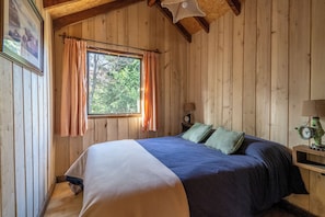 Ein Schlafzimmer einer Holzhütte mit einem Bett, einem Nachttisch, einer Uhr und einem Fenster mit Vorhängen.