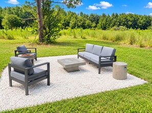 An outdoor seating area with a sofa, chairs, and a coffee table, surrounded by a gravel area and grass, with a forest in the background. - Foley Retreat | 4 Bedrooms • Grill & Smoker (Foley)