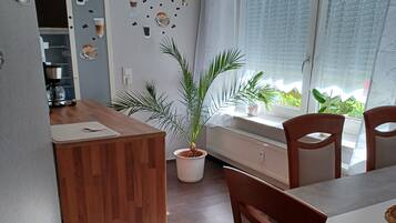 A dining area with a wooden table, chairs, a potted plant, and a kitchen visible in the background.
