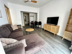 Een moderne woonkamer met een bank, een televisie op een houten kast, een eethoek met stoelen en een tafel, en een plafondventilator.