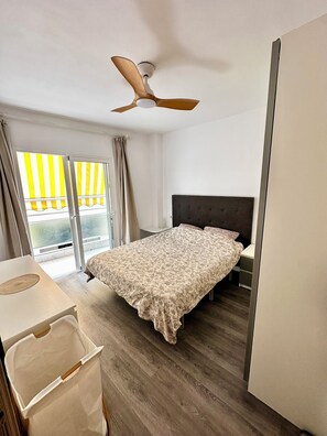 A bedroom with a bed, a ceiling fan, a window with curtains, and a view of a yellow building outside. - Great 2BR Apartment with 4 min walk everywhere (Playa San Juan)
