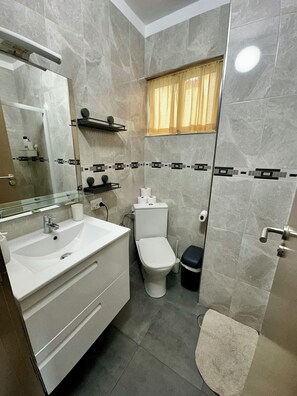 Een moderne badkamer met een witte wasbak, spiegel en toilet. Er is een raam met gordijnen, een handdoekhouder en een prullenbak.