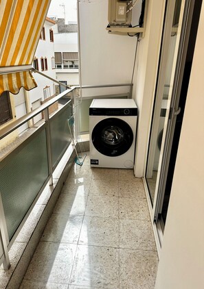 Een klein balkon met een wasmachine, een metalen hekwerk en uitzicht op gebouwen.