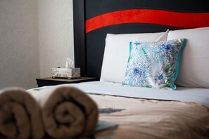 A neatly made bed with a floral pillow, a tissue box on a bedside table, and rolled towels in the foreground. - Casa Inn Retreat | Villas Morelos II (Puerto Morelos)