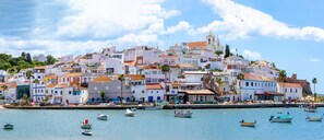 A coastal town with buildings along the waterfront, boats docked, and a prominent church on a hill. - Casa Eden (Ferragudo)