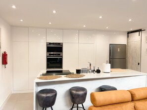 A modern kitchen with white cabinetry, a central island with stools, a stainless steel refrigerator, and a fire extinguisher on the wall. - Casa Eden (Ferragudo)
