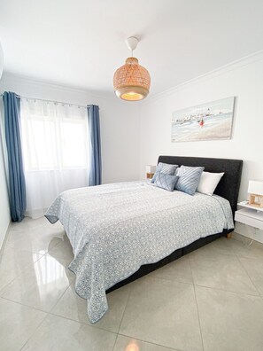 A bedroom with a bed, a nightstand, a lamp, a window with curtains, and a wall-mounted artwork. - Casa Joao (Carvoeiro)