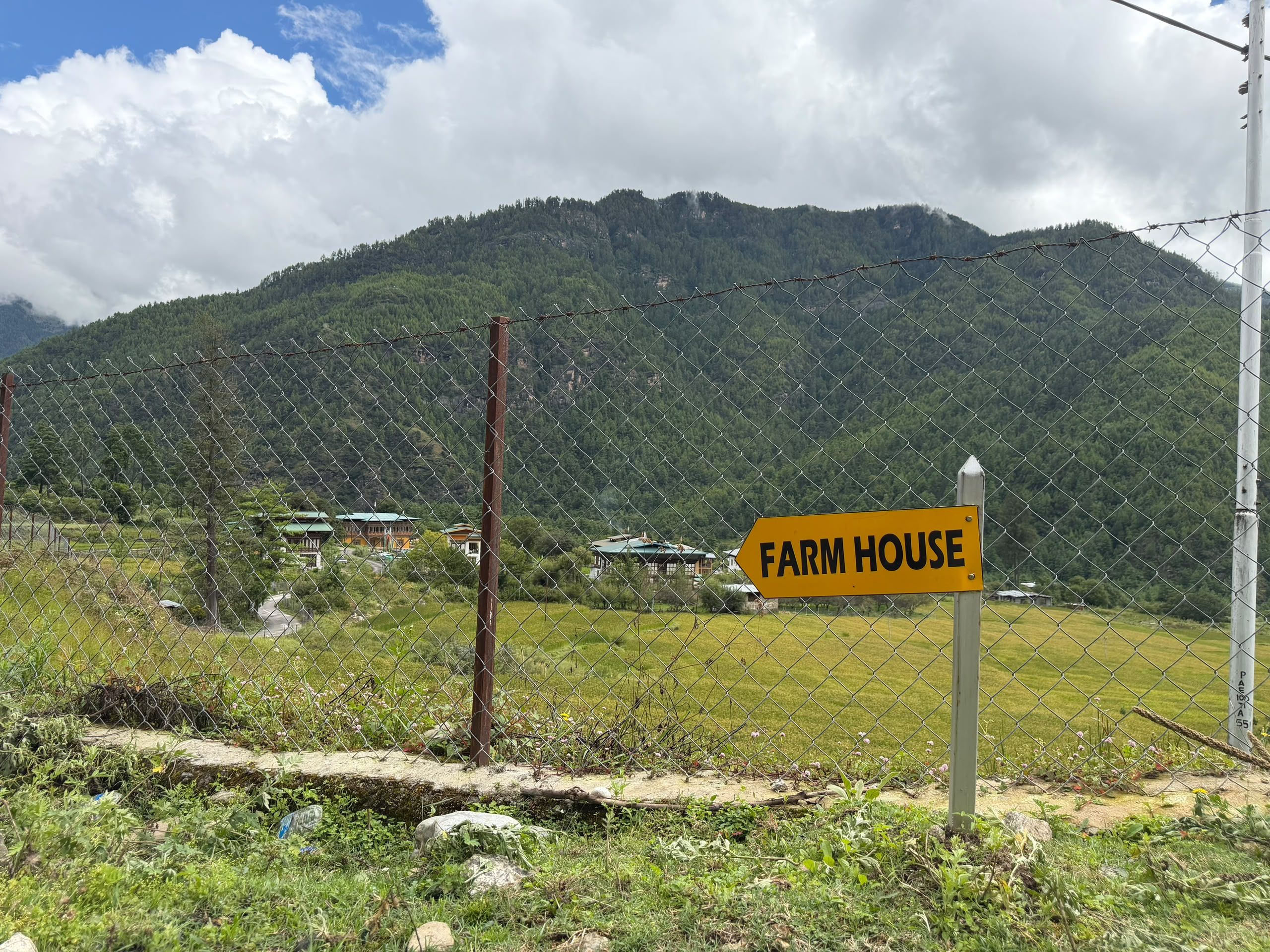 A fenced area with a sign indicating a farm house, surrounded by greenery and a mountain backdrop.
