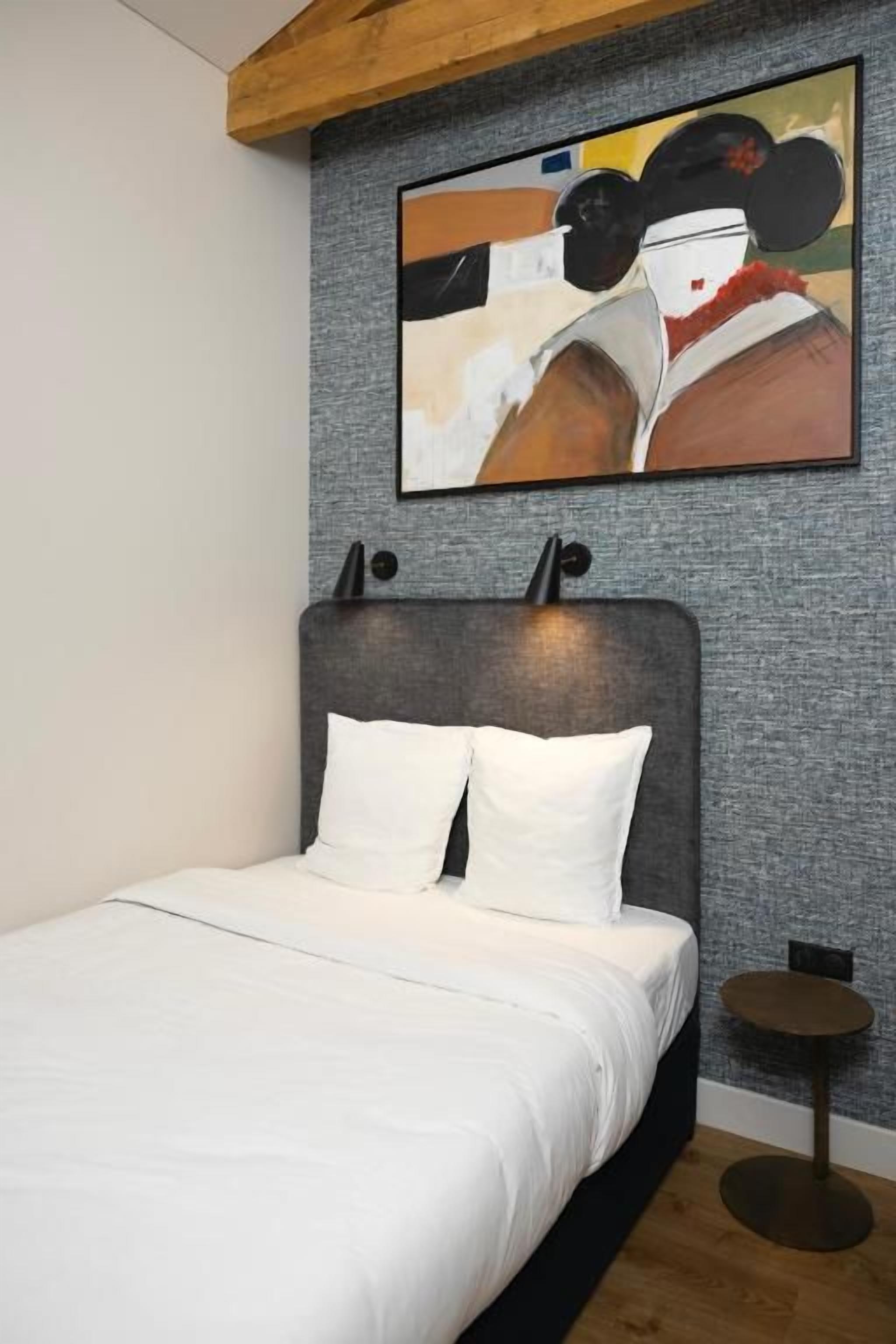 Un dormitorio con cama, cabecera, un cuadro en la pared y una mesita redonda.
