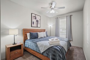 A bedroom with a bed, bedside table, lamp, ceiling fan, window with curtains, and a wall art piece. - Double T Rally House - 3BR/3BA - Walk to Tech (Lubbock)