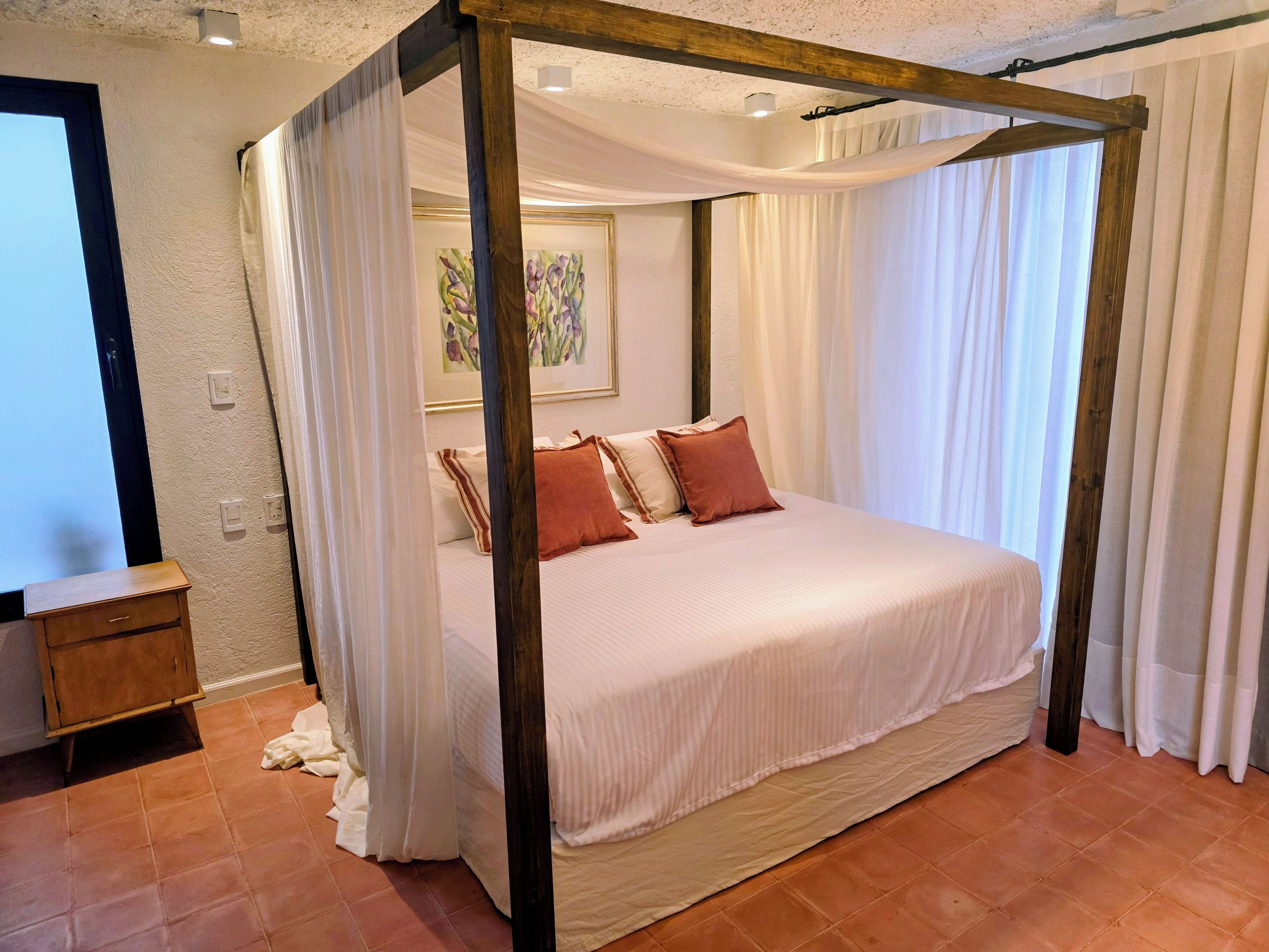 A four-poster bed with white bedding and red pillows, a wooden nightstand, and a painting on the wall.