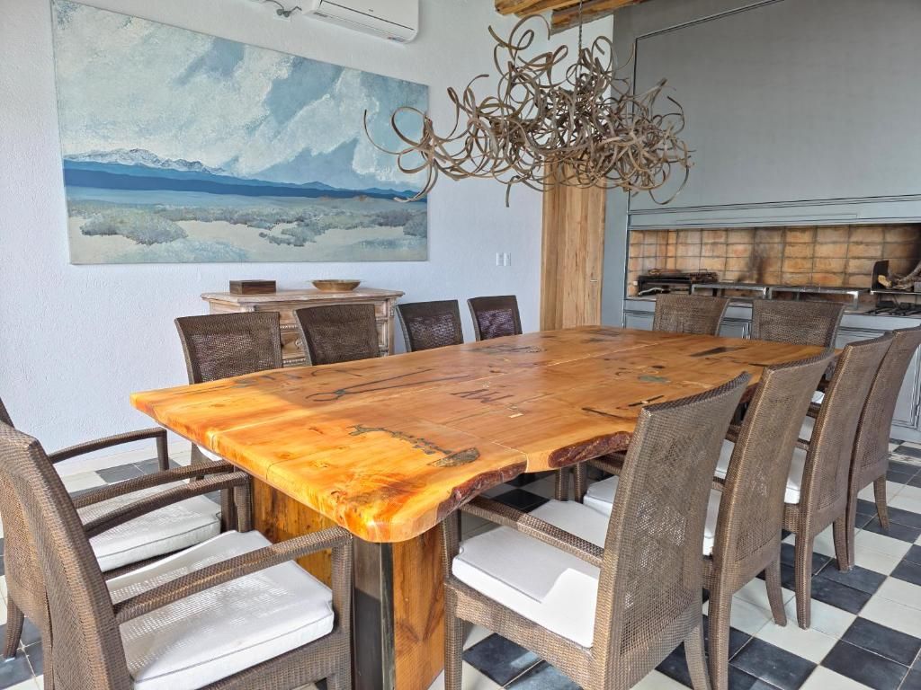 Un comedor con una gran mesa de madera rodeada de sillas de mimbre, un cuadro en la pared y una lámpara de araña moderna.