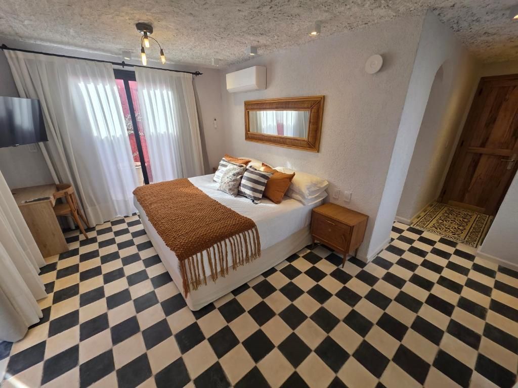 A hotel room with a checkered floor, a bed with a brown throw, a bedside table, and a mirror on the wall.
