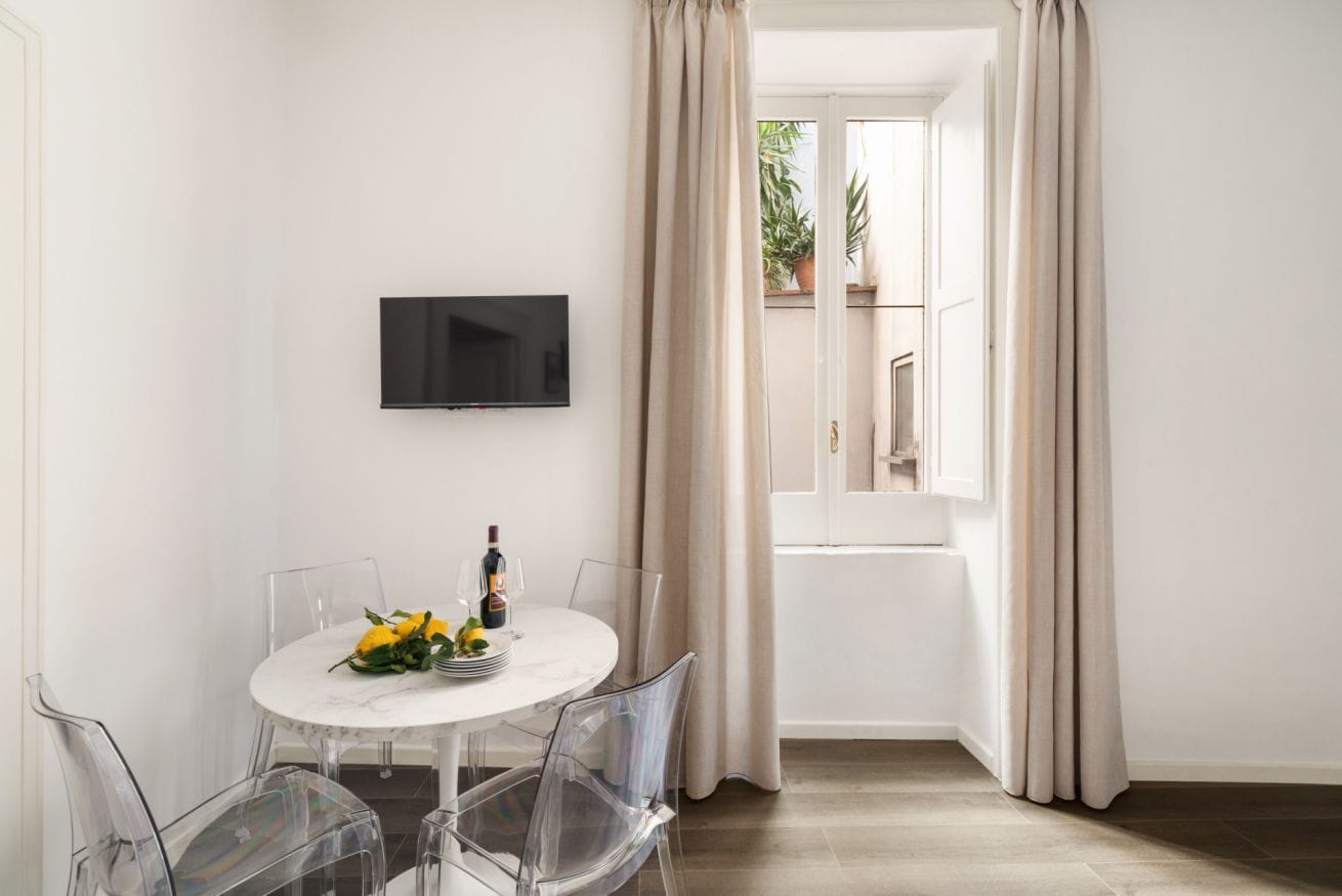 Un comedor con una mesa redonda, sillas transparentes, un televisor de pantalla plana montado en la pared y vistas a través de una ventana abierta.