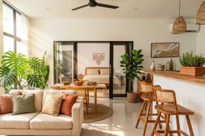 A modern living room with a sofa, a coffee table, and a dining area with chairs. There is a large window providing natural light, and the room has a ceiling fan. - Bohemian Luxury 3 BR  In the Center of Canggu with Pool (Canggu, Kabupaten Badung)