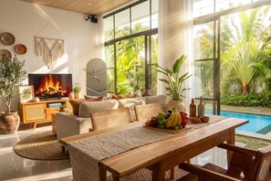 Un saló modern amb una llar de foc, seients confortables, una zona de menjador amb taula preparada per a dos, i un gran finestral amb vista a un jardí exuberant i piscina.