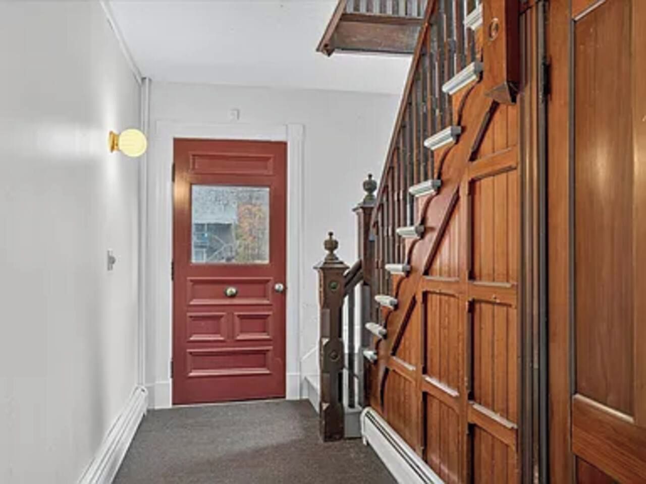 A hallway with a red door, a wooden staircase, and a wall-mounted light fixture.
