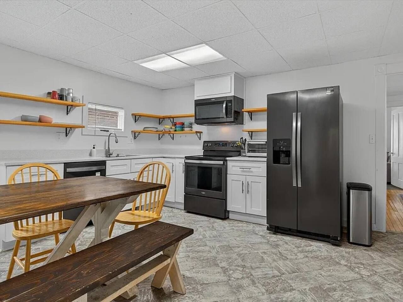 A modern kitchen with white cabinets, a stainless steel refrigerator, a microwave, and a stove. There is a wooden dining table with chairs.