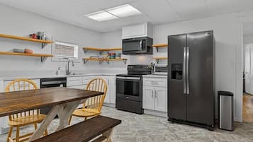 A modern kitchen with white cabinets, a stainless steel refrigerator, a microwave, and a stove. There is a wooden dining table with chairs.