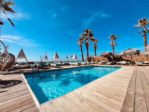 A wooden deck with a pool, lounge chairs, and palm trees. - Athena Seafront Apartment Parking Pool (Castel Volturno)