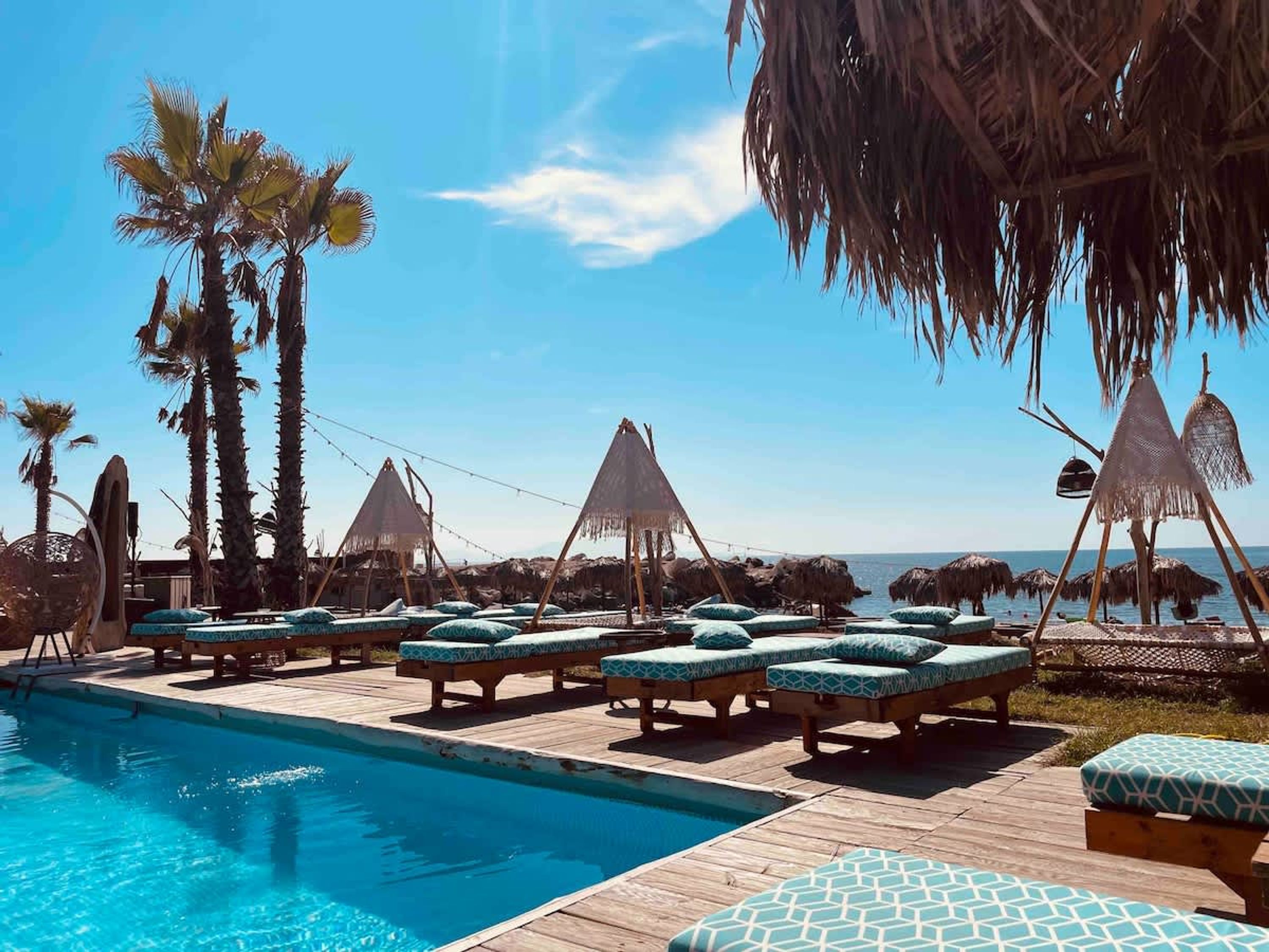 A poolside area with lounge chairs and thatched umbrellas, palm trees, and a view of the sea.