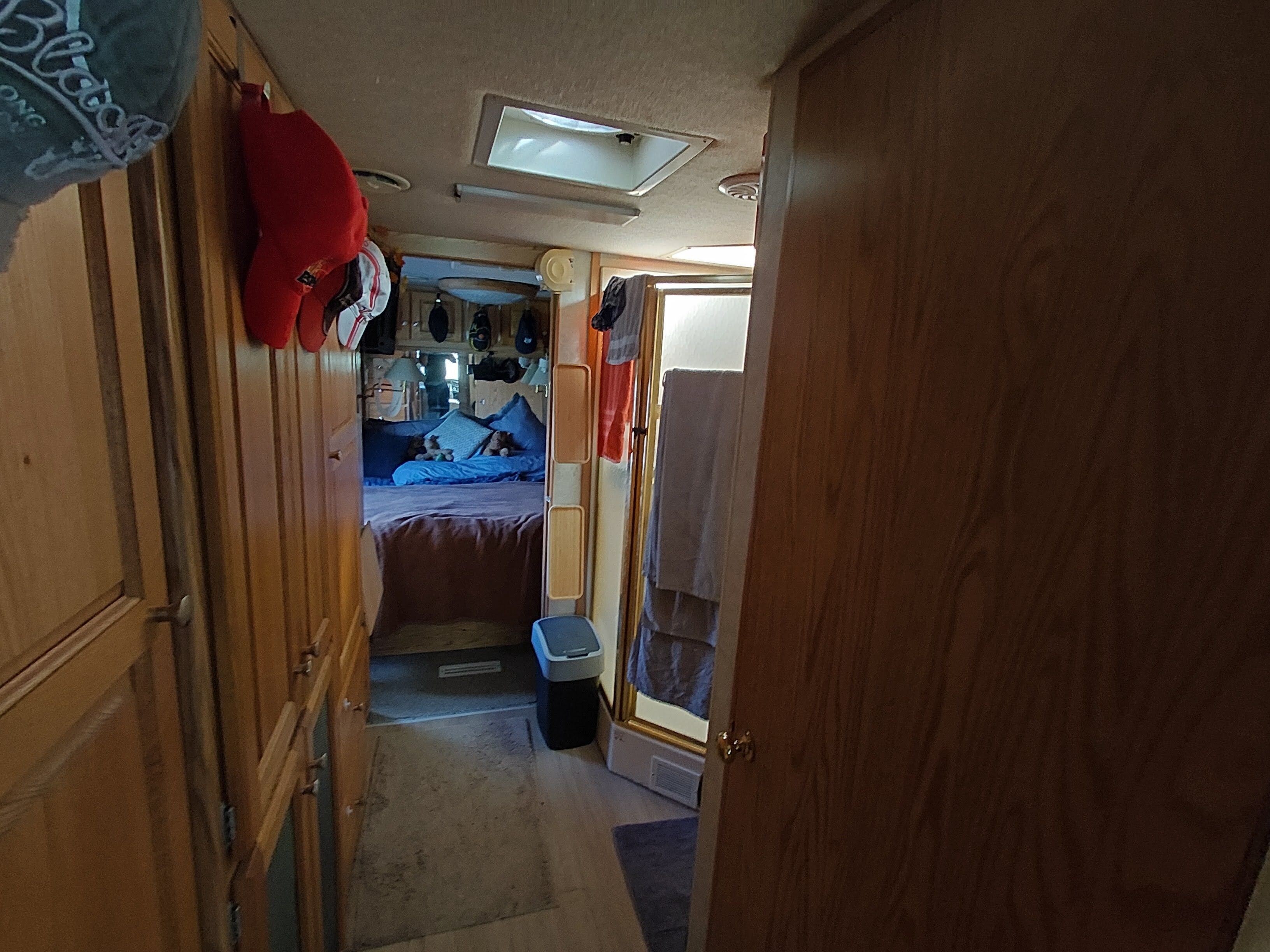 Ein schmaler Gang führt zu einem Schlafzimmer mit einem Bett, einer kleinen Mülltonne und einer Tür zu einem weiteren Raum.