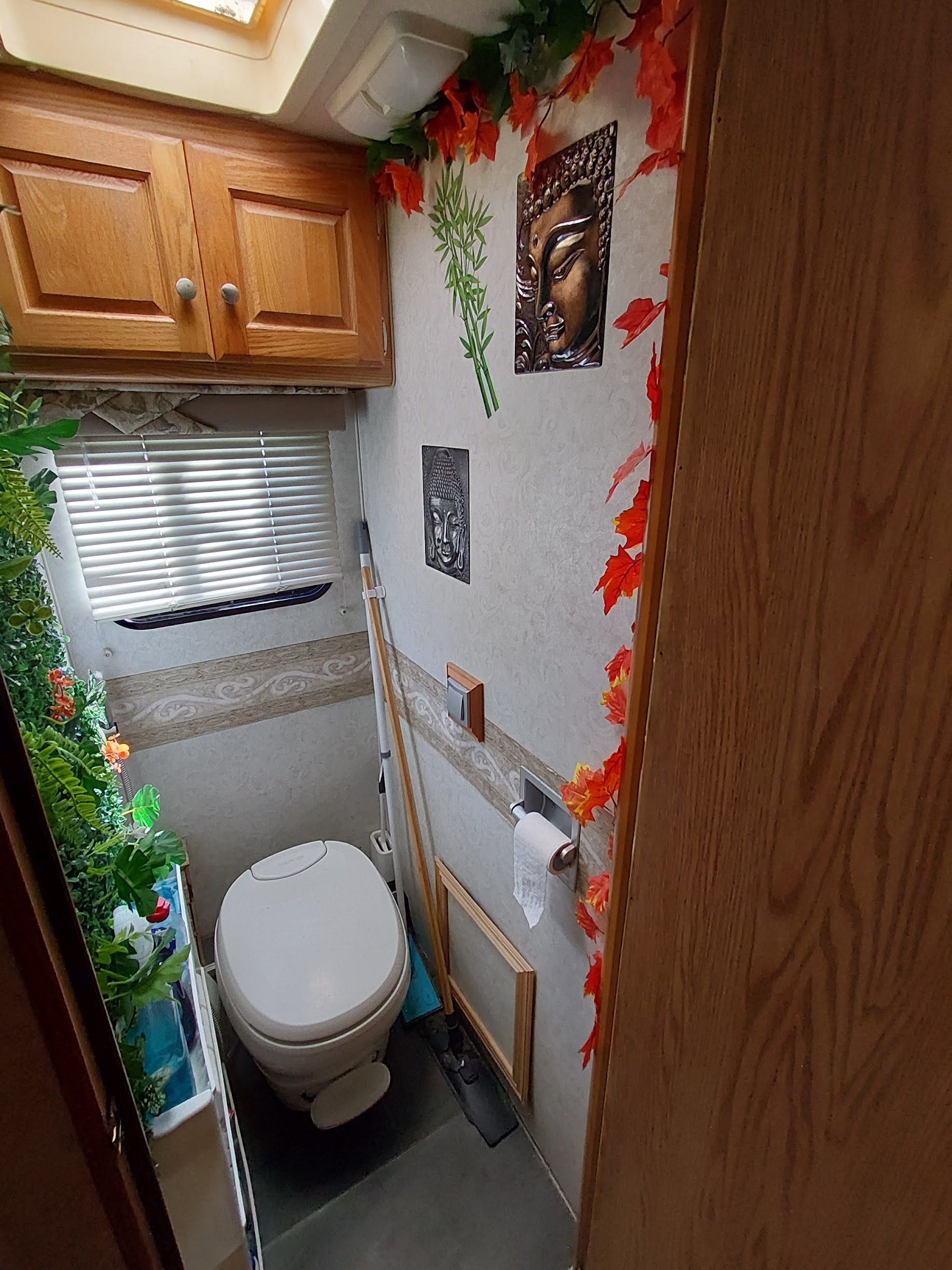Un baño pequeño con inodoro, una ventana con persianas y decoración en la pared que incluye imágenes de Buda y hojas otoñales.