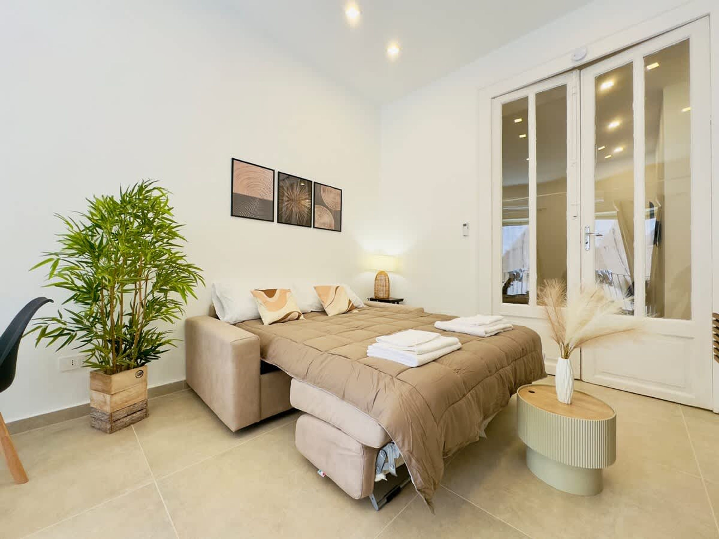 Un dormitorio moderno con sofá cama, mesita de noche con lámpara, ventana grande y planta en maceta.