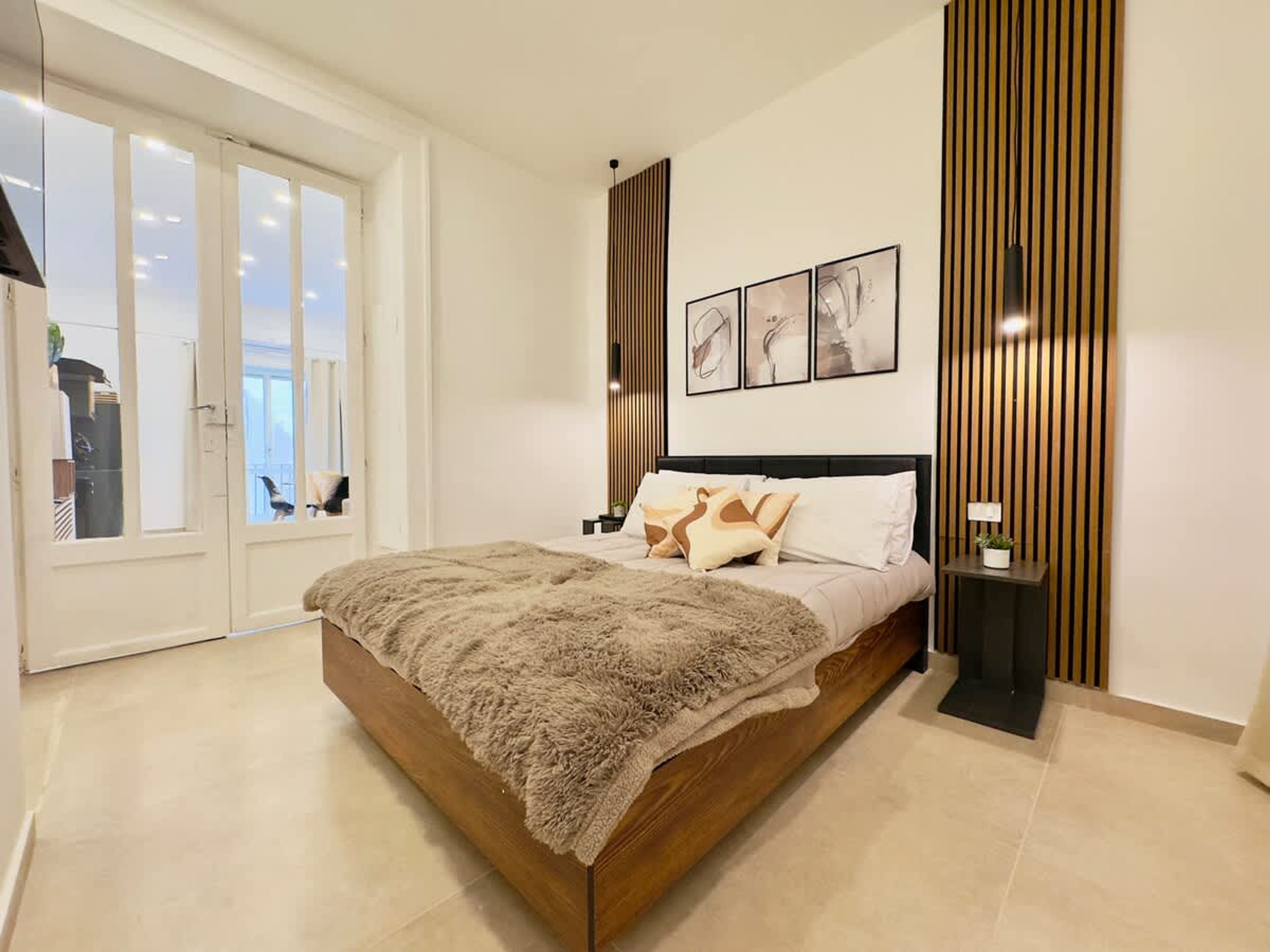 Un dormitorio con una cama grande, una mesita de noche, una lámpara de pared y una puerta corredera que da a un balcón.