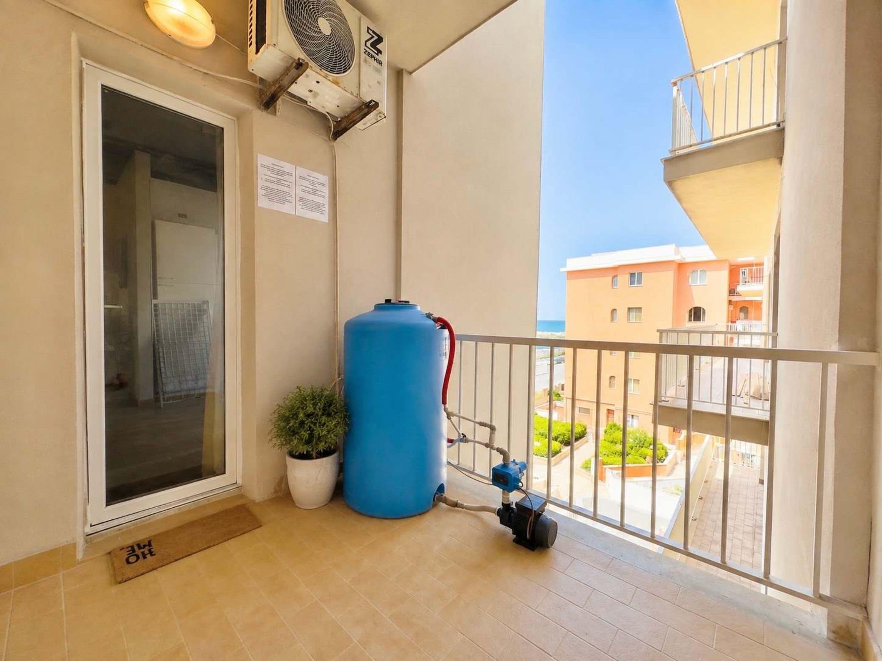 Un balcón con un tanque de agua azul, un pequeño ventilador y una planta en maceta.