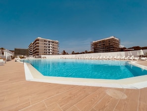 Una piscina con sillas de descanso y sombrillas, rodeada de edificios de varios pisos.