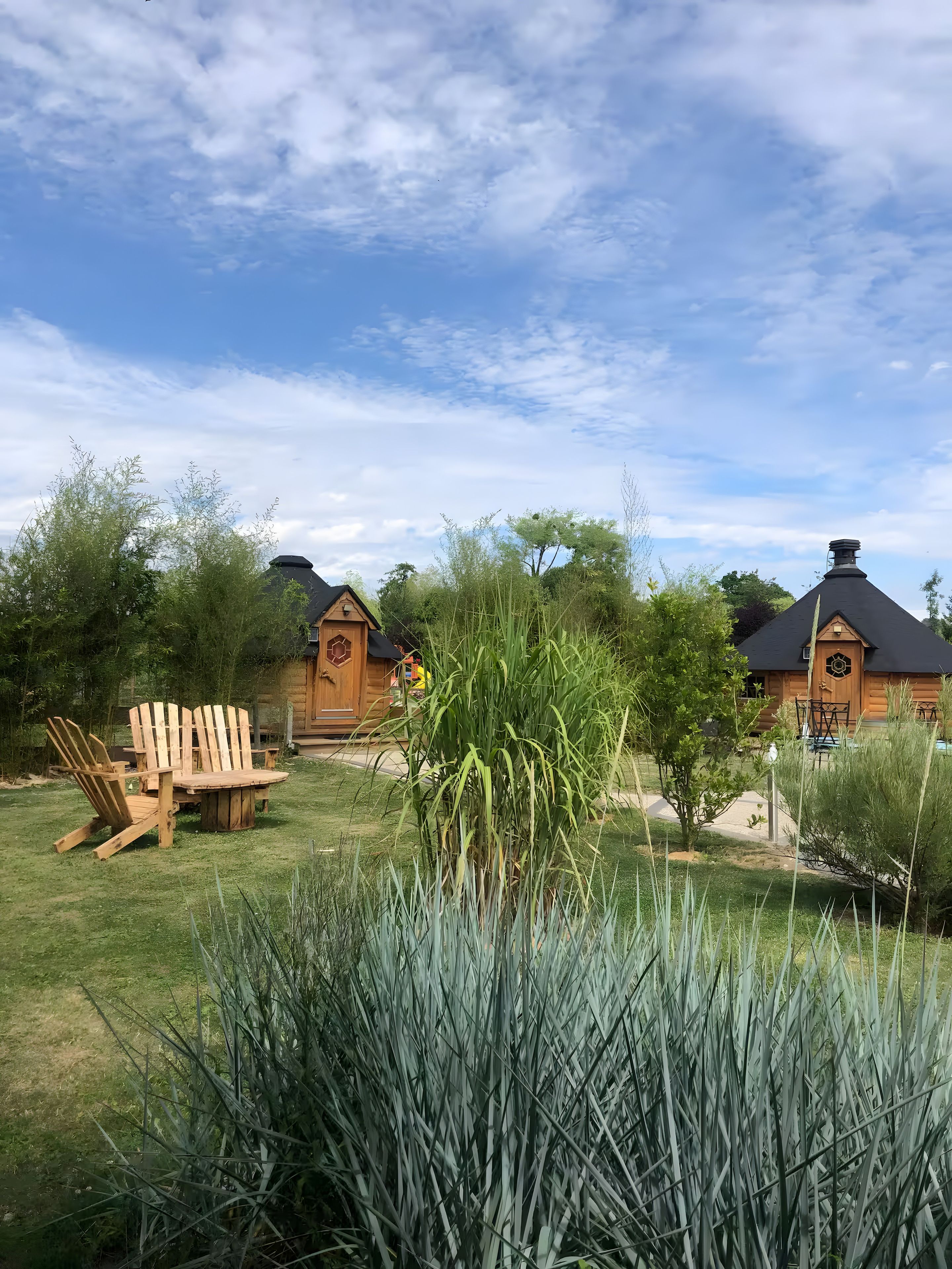 A wooden cabin with a round table and chairs in a grassy area, surrounded by tall grasses and trees.