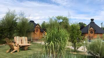 A wooden cabin with a round table and chairs in a grassy area, surrounded by tall grasses and trees.